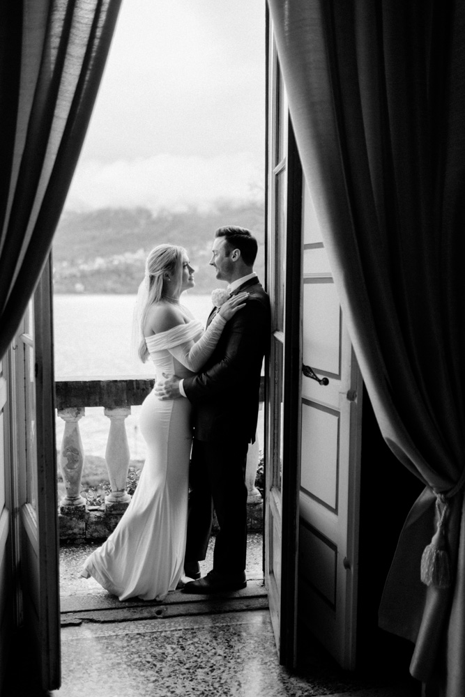 A black and white picture for the couple holding their hands on the balcony over the Como lake, framed in the window