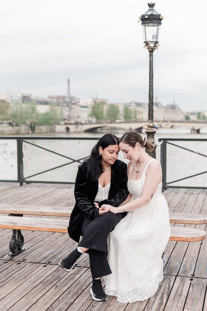 The 2 brides sit on a bench at Pont des Arts in paris